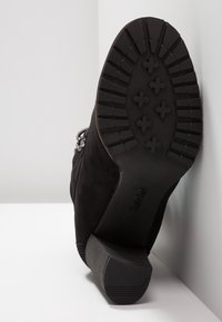 Bottine en daim noir avec une semelle en caoutchouc texturée, un talon bloc et des ornements décoratifs sur le côté.