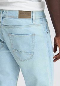 Jeans de mezclilla azul claro con una etiqueta de cuero, que presentan bolsillos traseros en ángulo y costuras amarillas. El diseño es de corte relajado con una textura suave.