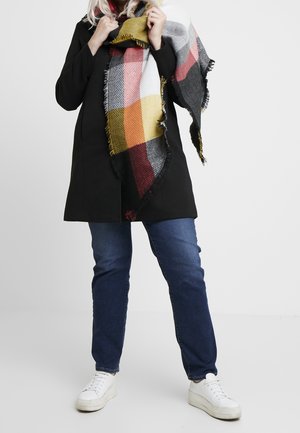 Personne portant un manteau sombre, un jean bleu, des baskets blanches et une grande écharpe à carreaux multicolores enroulée autour du cou.