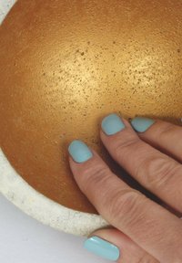 Plat rond et texturé avec un intérieur doré et un extérieur en béton blanc. Une main avec des ongles bleu clair repose légèrement sur le plat.