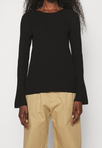 Pull en maille côtelée noir à manches longues et col rond large, avec des poignets évasés et un ourlet légèrement allongé.