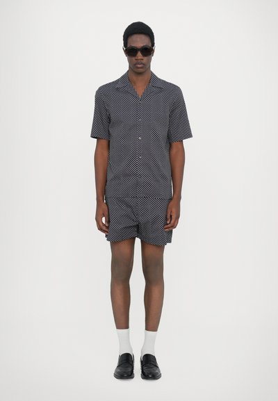 Chemise à manches courtes à pois noir et blanc et shorts assortis, fabriqués en tissu léger, associés à des chaussures noires et des chaussettes blanches montantes.