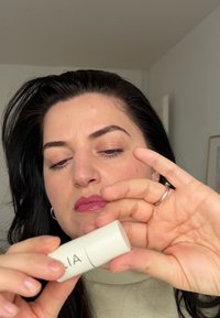 Ein zylindrischer, weißer Kosmetikstift mit der Aufschrift "ILIA", der in einer Hand gehalten wird. Das Produkt hat eine glatte Textur und der Hintergrund ist weich und neutral.