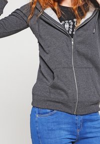 Felpa grigia con zip e cappuccio foderato di grigio chiaro, tasche frontali e polsini a coste. Indossata con jeans blu e una t-shirt nera con grafica sotto.