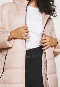 Kvinna i en ljusrosa Nike-puffjacka över en vit skjorta och svarta byxor, som justerar dragkedjan på jackan med båda händerna.