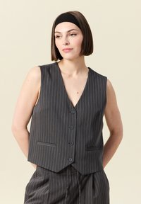 Grå väst med smalrandig mönster, med notch-krage, fem knappar och två framfickor. Tyget har en slät textur som kompletterar skräddarsydda byxor.