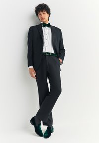 Smoking nero con rever a punta in raso, camicia bianca, fiocco e cintura in velluto verde, pantaloni neri e scarpe in velluto verde scuro.