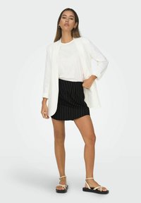 Witte op maat gemaakte blazer over een plain witte t-shirt, gecombineerd met een zwarte minirok met streepjes en lichte sandalen met een zwarte zool.