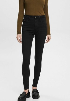 Kobieta w czarnych obcisłych dżinsach, brązowej bluzce z długim rękawem i czarnych mokasynach, stojąca na jednolitym jasnym tle.