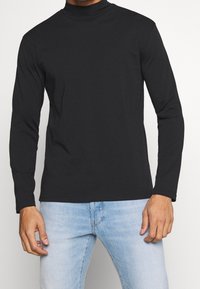 Schwarzes Langarm-Turtleneck-Shirt aus glattem Stoff, mit figurnahem Schnitt und schlichter Verarbeitung. Kombiniert mit hellblauen Jeans.