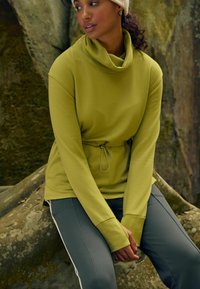 Maslinastosiva dug rukav sweatshirt s krojem s kapuljačom i podesivim kaišem u struku. Uparen s asosijativnim sivim sportskim hlačama s bočnim trakama.