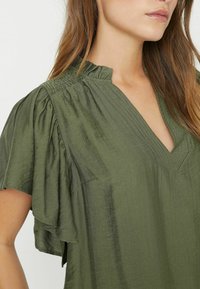 Olivgrüne Bluse mit V-Ausschnitt, gerafften Schultern und kurzen, rüschierten Ärmeln. Der Stoff wirkt leicht und hat eine subtile Textur.