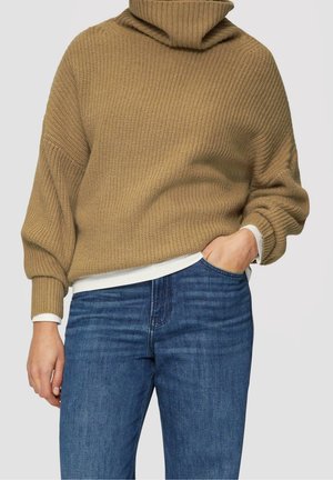 Personne portant un pull à col roulé marron en tricot à manches longues par-dessus une chemise blanche et un jean bleu, main dans la poche.