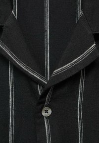 Zwarte shirt met witte verticale strepen, gemaakt van gestructureerde stof. Heeft een opgerolde kraag en een enkele grijze knop aan de voorkant.