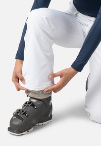 White ski pants with a cuff adjustment feature are worn over a black ski boot. The pant legs are fitted, with a smooth fabric texture.