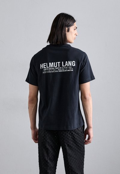 T-shirt in cotone nero con il nome del marchio "HELMUT LANG" e testo bianco sul retro. Il modello indossa pantaloni neri con texture.