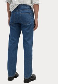 Jeans de mezclilla azul oscuro con un corte recto, que cuentan con dos bolsillos traseros y detalles de costura mínimos, combinados con zapatos negros.