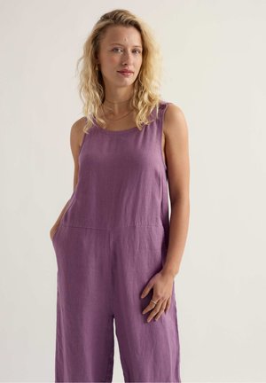 Laineliste blondide juustega naine, kes kannab varrukateta lillat kombekat taskutega, seisab ühetoonilise heleda tausta ees.