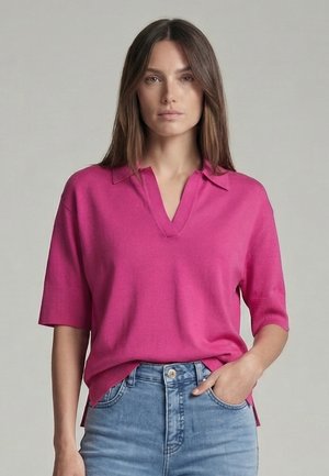 Frau mit langem braunem Haar trägt ein knallpinkes Poloshirt mit kurzen Ärmeln und hellblaue Jeans, steht vor einem schlichten grauen Hintergrund.