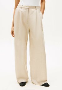 Pantaloni beige a gamba larga con pieghe davanti, due tasche laterali e una texture liscia. Abbinati a mocassini neri per un contrasto.