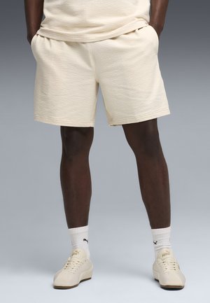 Homme portant un short beige texturé, des chaussettes blanches Puma, et des baskets beige Puma, debout les mains dans les poches devant un fond uni.