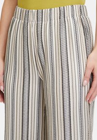 Gestreepte wijde broek met een elastische tailleband in crème, zwart en beige. De textuur van de stof heeft verticale strepen en decoratieve stiksels.