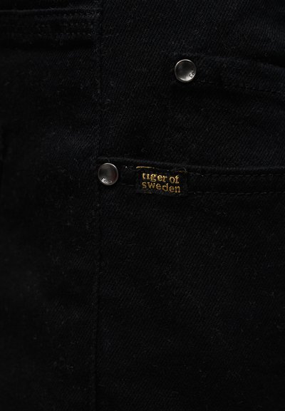 Mustast teksariidest kangas, millel on metallist naelad ja väike must kangast silt kollase tikkega, millel on kiri "tiger of sweden".