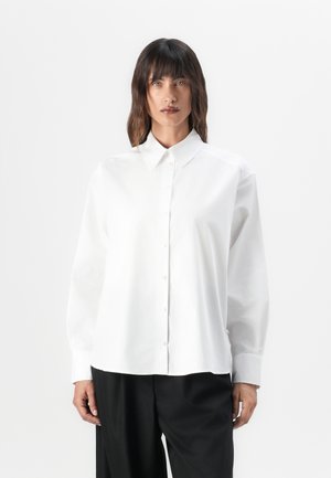 Camisa branca de mangas longas com colarinho pontiagudo, feita de tecido de algodão suave, apresentando um corte descontraído e costuras de ombro discretas.
