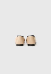 Scarpe in pelle beige con accenti neri, suole piatte e design arrotondato. Presentano un piccolo logo sul tallone per l'identificazione del marchio.