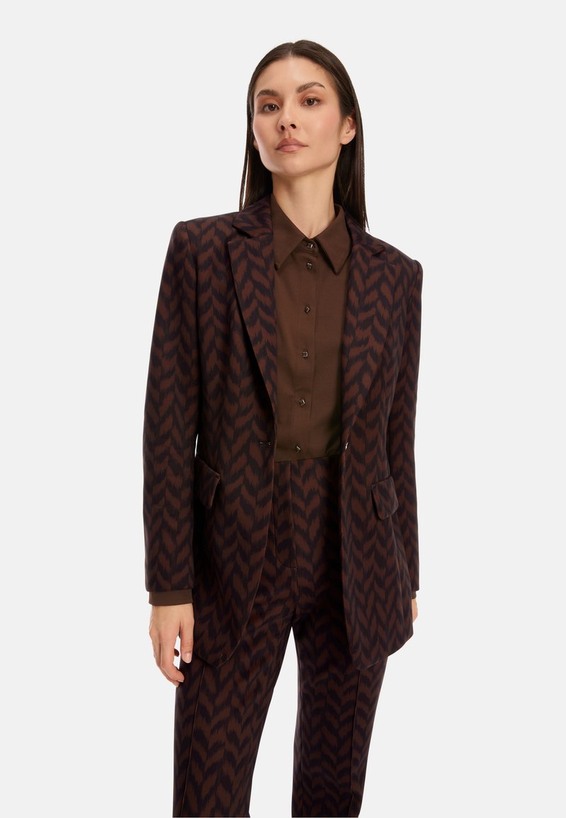 Brown patterned blazer with matching trousers, featuring sharp lapels and front pockets, worn over a brown button-up shirt. Smooth texture.