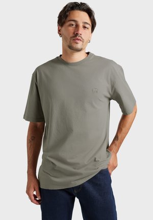 Homme aux cheveux courts et foncés et portant des boucles d'oreilles, vêtu d'un t-shirt gris à manches courtes et d'un jean bleu foncé, debout avec une main dans la poche.