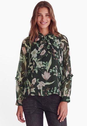 Femme portant un chemisier noir transparent avec un imprimé floral vert et rose et un col à nouer, associé à un jean foncé, souriant à la caméra.