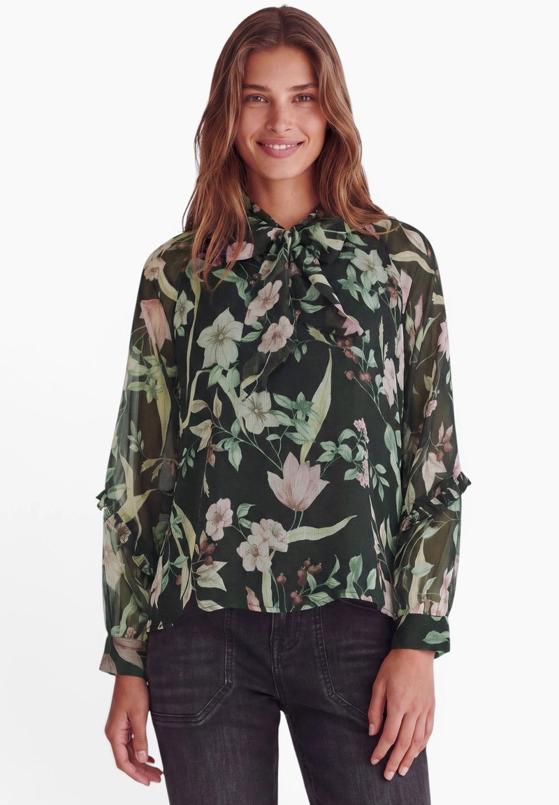 Femme portant un chemisier noir transparent avec un imprimé floral vert et rose et un col à nouer, associé à un jean foncé, souriant à la caméra.