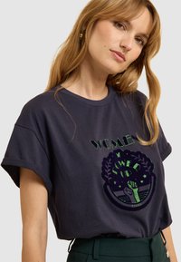 T-shirt en coton bleu marine avec des manches roulées, arborant un graphisme métallique vert d'un poing levé et du texte "WOMEN POWER UP", entouré de motifs de feuilles.