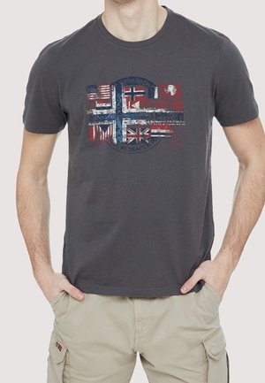 Homme portant un t-shirt gris foncé avec un motif usé représentant plusieurs drapeaux sur un emblème circulaire bleu, associé à un short cargo beige.