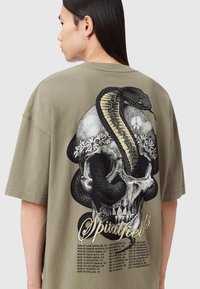 Olivengrøn oversized t-shirt med et kraniegrafik, der inkluderer en slange og blomsterdetaljer på ryggen, med tekst og turnédatoer nedenunder.