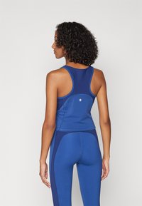 Kék sportos trikó, versenyhátra kialakítással, sima textúrával és diszkrét díszítéssel, kék leggings-el párosítva.