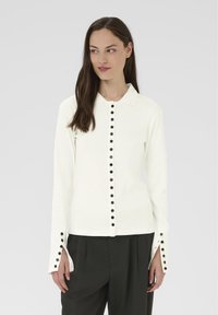 Kobieta ubrana w białą bluzkę z długim rękawem, z czarnymi guzikami z przodu i przy mankietach, w połączeniu z czarnymi spodniami z plisami, na szarym tle.