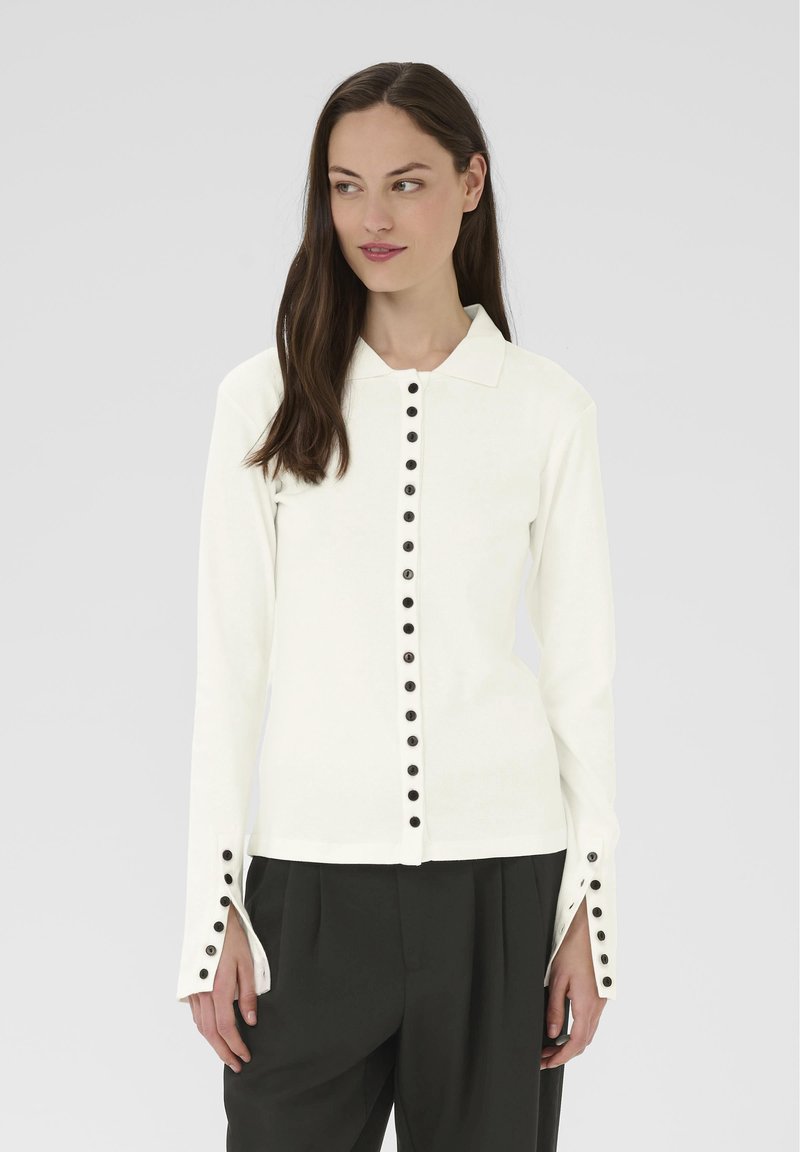 Kobieta ubrana w białą bluzkę z długim rękawem, z czarnymi guzikami z przodu i przy mankietach, w połączeniu z czarnymi spodniami z plisami, na szarym tle.