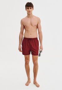 Czerwone kąpielówki z elastycznym paskiem w talii, wykonane z lekkiego materiału. Model trzyma w ręku okulary przeciwsłoneczne i stoi boso, pozując wprost do obiektywu.