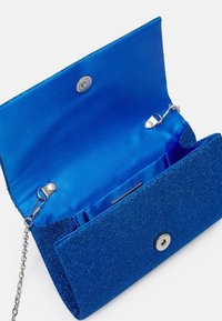 Blå glittrig kuvertväska med satingolv, metallknappslutning och avtagbar silverkedjeaxelrem mot vit bakgrund.