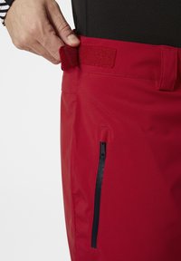 Červené voděodolné kalhoty s páskem na suchý zip, mají boční zipovou kapsu s černým zipem a hladkou, matnou texturu.