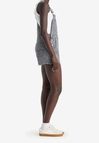 Graue Jeans-Doverallkleid mit verstellbaren Trägern, Fronttaschen und fransigem Saum, kombiniert mit weißen Sneakers und Knöchelsocken.