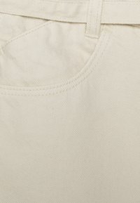 Tejido de algodón beige con un bolsillo de costura curva, textura suave y una cinturilla visible con pasadores para el cinturón.