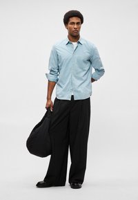 Ανοιχτό μπλε πουκάμισο με σχέδιο, με μακριά μανίκια, συνδυασμένο με χαλαρές μαύρες παντελόνες. Το μοντέλο κρατά μια μαύρη τσάντα με καπιτονέ σχέδιο.