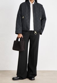 Persoon draagt een gewatteerde zwarte jas, wit overhemd, wijde zwarte broek, zwarte schoenen, houdt een kleine gestructureerde zwarte handtas in de linkerhand.