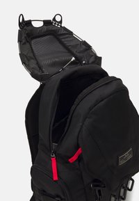 Black backpack with a textured exterior, featuring a zippered opening, red zipper pull, and a mesh lining inside.
