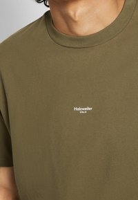 Gros plan d'une personne portant un t-shirt vert olive avec un petit logo blanc « Holzweiler Oslo » sur le côté gauche de la poitrine.