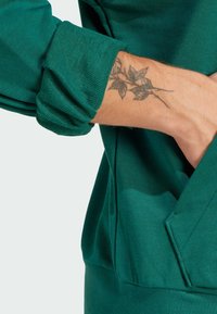 Zelená mikina s štruktúrovanou látkou, prehnutými manžetami a klokany vakom. Obsahuje tetovanie lístia na predlaktí.