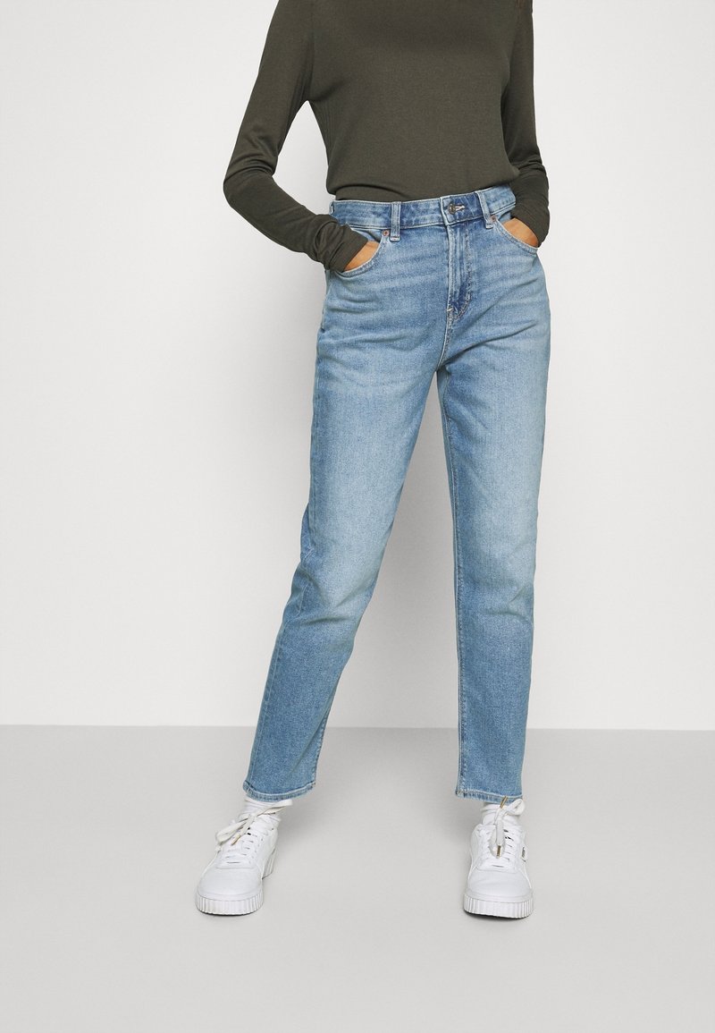 Personne portant un jean taille haute bleu clair, un haut à manches longues vert foncé, les mains dans les poches et des baskets blanches sur un fond uni.